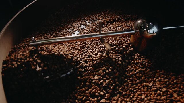 Tracking shot of roasted coffee beans tumbling and stirred inside industrial roaster drum in roastery.