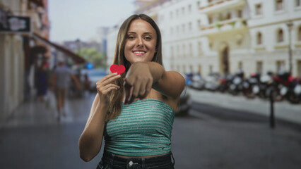 Fototapeta premium Young woman holding a small red paper heart and pointing her finger at the viewer on a busy city street lined with scooters and pale building facades; playful romance connection.