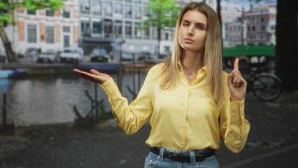 Fototapeta premium Woman holds palm up and points index finger on street by a canal, eyes closed in thoughtful pose; contemplation.