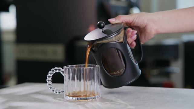 Closeup of hand pouring french press coffee into glass mug for menu design on background roasting shop.