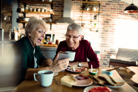 Senior women friends at breakfast, smiling over smartphone in cozy home kitchen