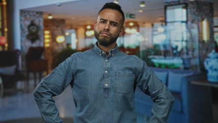 Naklejka premium Man with arms akimbo in hotel lobby near reception desk wearing denim shirt and frowning at camera; doubt questioning.