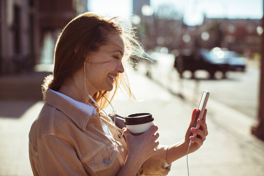 Smiling young woman using smartphone and earphones on city street