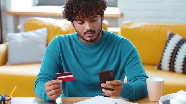A young man with curly hair looks at his phone while holding a credit card. He appears focused on an online shopping task. The setting is a cozy living room with a yellow sofa.