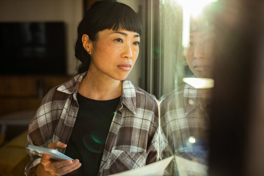 Thoughtful woman with smartphone looking out home window