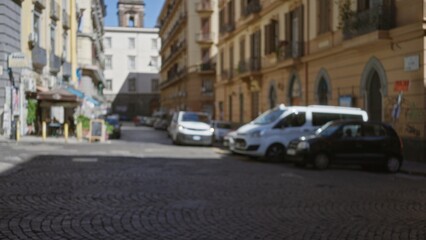Fototapeta premium Narrow cobblestone street with parked vans and blurred building facades, soft defocused bokeh background; backdrop, copyspace, backplate, calm.