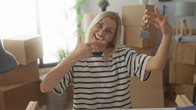 Woman holding house keys and points finger at key amid stacked moving boxes in a building, smiling broadly and showing hand gesture; joy new home.