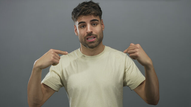Man wearing casual beige short sleeve t shirt points both index fingers to chest in gray studio; confidence.
