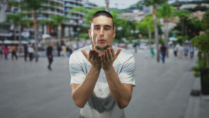 Fototapeta premium Man blows kiss with cupped hands to mouth on crowded urban street plaza, wet face with water droplets and casual striped tshirt; affection warmth.