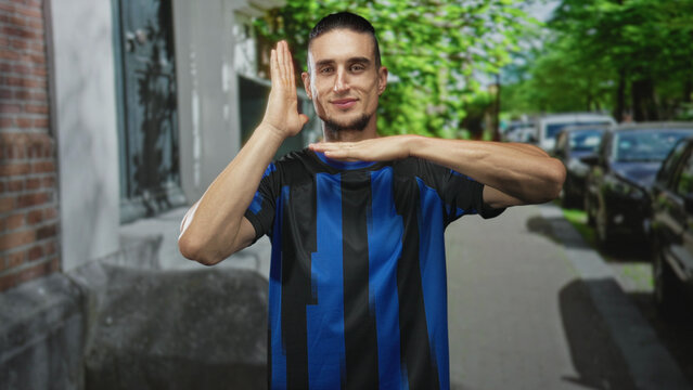 Man in blue and black soccer jersey forms timeout hand gesture with visible hands and forearms on a leafy street with parked cars and brick building; team pride confidence.