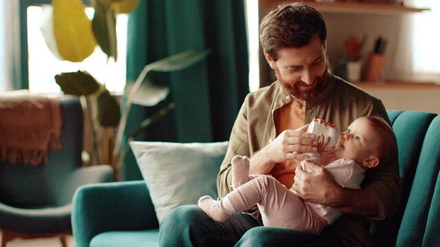 A man with a beard sits in a comfortable chair while feeding a baby with a bottle. The room is bright and has plants and soft furniture in the background, creating a warm atmosphere.