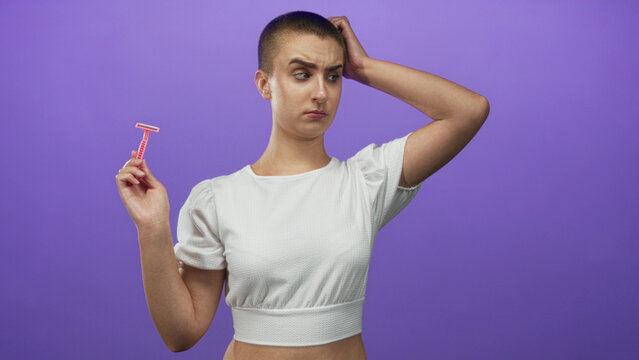 Woman with buzz cut holding pink disposable razor and hand on head in studio with purple backdrop wearing white crop top and puzzled expression; hesitation doubt.