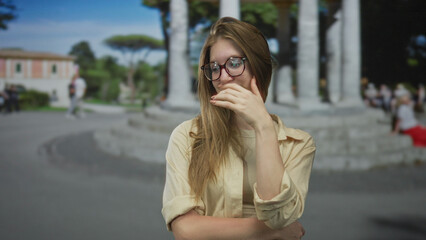 Obraz premium Young blonde woman wearing beige shirt adjusting glasses and touching cheek on street; embarrassment.