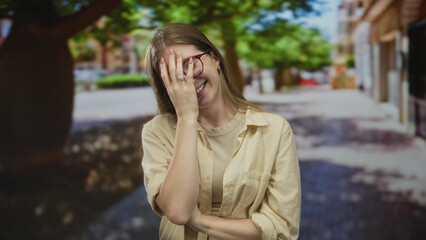 Fototapeta premium Young blonde woman wearing glasses and a light shirt covers mouth with hand on bustling city street; amusement.