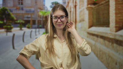 Obraz premium Blonde woman adjusts glasses with hand touching ear by a brick building on street; quiet confidence.
