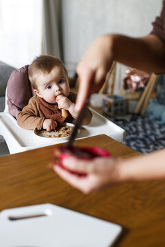 Mother feeding cute baby infant with fuit in high chair, eating complementary food. Family domestic life. Mom and son together, joyful moment.