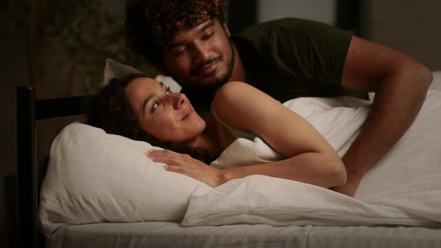 A man with dark skin and a woman with light skin embrace and smile while lying in bed. They share a joyful moment wrapped in white sheets in their cozy bedroom.