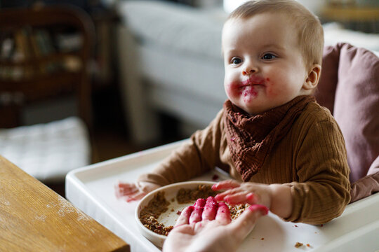 Mother feeding cute baby infant with fuit close up in high chair, eating complementary food. Family domestic life. Mom and son together, joyful moment.