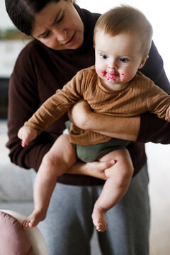 Mother holding cute baby infant with messy face after feeding complementary food. Family domestic life. Mom and son together, joyful moment. Happy Mother's day!