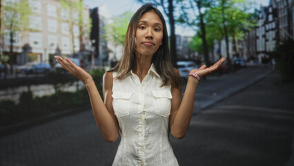 Thai woman stands facing camera with hands raised in a shrug and pointing gesture on a city street lined with cars and buildings  quiet uncertainty. © Krakenimages.com