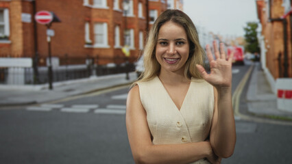 Fototapeta premium Woman waving right hand on a city street in front of brick buildings, palm visible and sleeveless beige vest worn; friendly greeting.