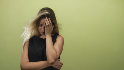 Fototapeta premium Woman with hand on forehead and folded arm, blonde wearing black sleeveless dress, long hair, looking down in studio with pale green wall; stress.