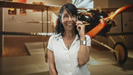 Woman talking on phone, smiling with phone to ear in a building beside a vintage biplane exhibit  joyful travel nostalgia. © Krakenimages.com