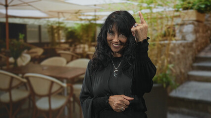 Fototapeta premium Senior hispanic woman shows middle finger with rings and smiling hand gesture in a building cafe patio with chairs and umbrellas; playful defiance.