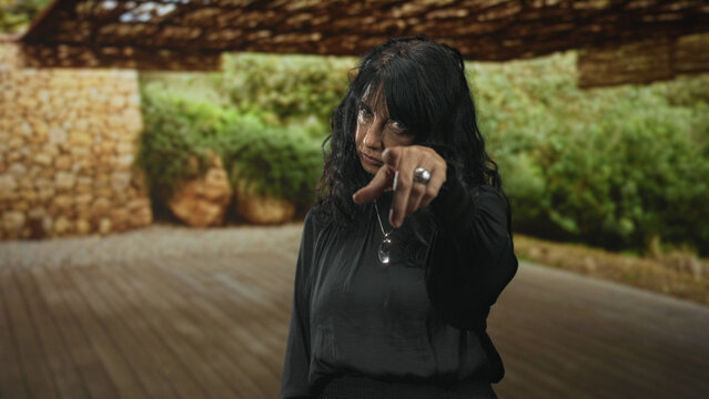 Woman pointing finger directly at camera on a wooden floor inside a building, wearing black blouse, pendant and ring; accusation authority.