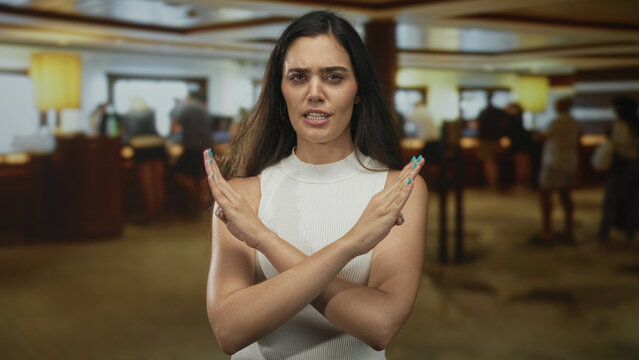 Young hispanic woman crosses arms and hands in denial at hotel lobby as eager guests wait behind; denial.