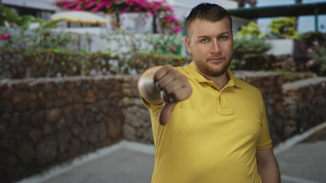Man, young caucasian in yellow polo showing thumbs down with outstretched hand on street near low rock wall and flower planters; disapproval.