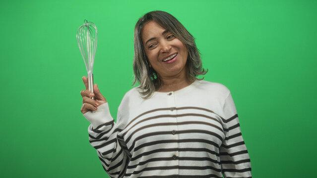 Hispanic woman holds whisk up and smiles, wearing striped sweater, posed before a studio green screen for cooking concept; joy.