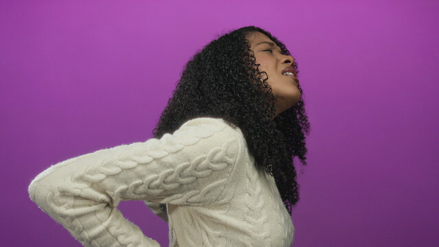 Woman grimaces while holding her lower back in a purple studio setting, showing pain and strain; discomfort.