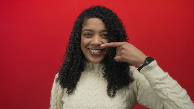 Smiling woman with curly hair wearing cream sweater points finger to mouth in studio with red wall; playfulness.