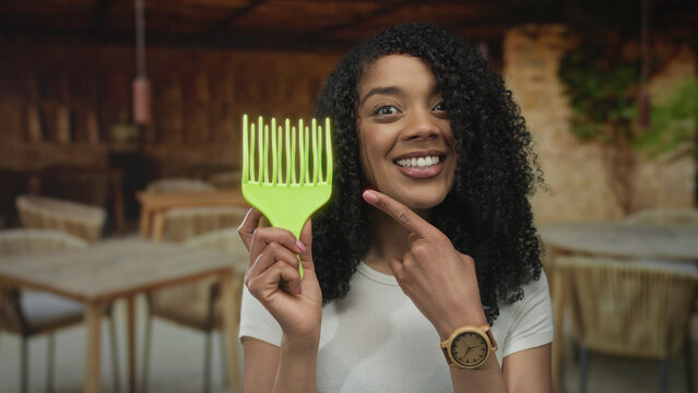 African american woman smiling points finger to green afro pick at outdoor restaurant terrace; confidence beauty empowerment selfesteem.