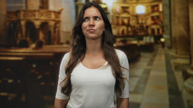 Woman frowning with furrowed brow inside catholic church interior, white shirt visible among pews and columns; doubt reflection faith.