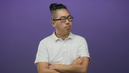 Fototapeta premium Man with crossed arms and glasses, wearing white polo and topknot, looking sideways in studio against a purple wall; skepticism introspection.