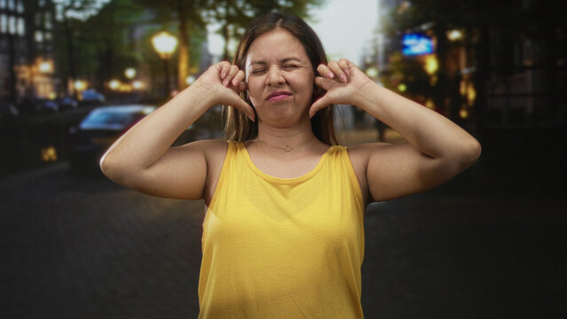 Woman plugs ears with fingers and eyes closed on busy city street at dusk wearing yellow tank top and necklace; annoyance urban noise.