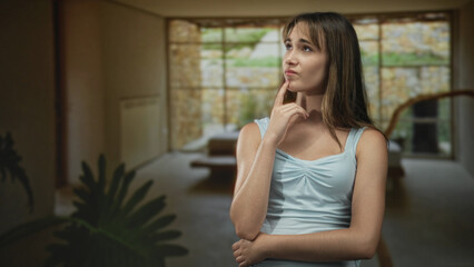 Fototapeta premium Young woman with finger to chin and folded arm in a living room inside a residential building; quiet contemplation.