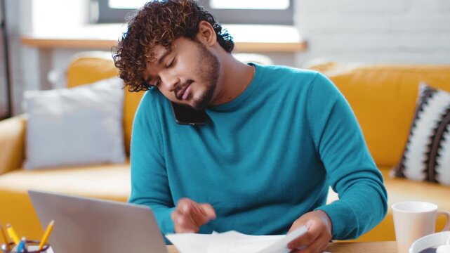 A man with curly hair is talking on his phone while typing on his laptop. He is seated on a sofa in a bright living room with a mug nearby and documents on the table.