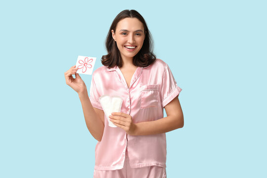 Beautiful young woman holding menstrual pads and paper with drawn flower on blue background. PMS concept