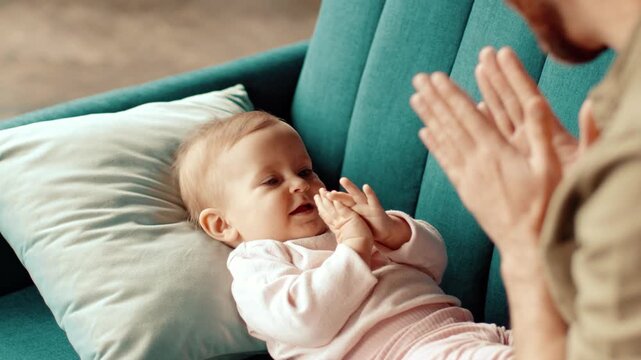 A man with light skin plays with his baby sitting on a couch. The baby looks curious and is reaching out towards the man's hands. The setting is warm and inviting, showcasing a cozy living space.