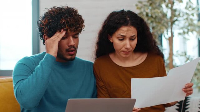 A man with curly hair and a woman with long dark hair sit together on a couch. They look focused as they review printed documents while using a laptop. It is midday in a stylish living room setting.
