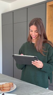 A cheerful young woman in a cozy green sweater smiles while scrolling on her digital tablet in a bright, contemporary kitchen setting. She appears relaxed and engaged with her device, perhaps enjoying