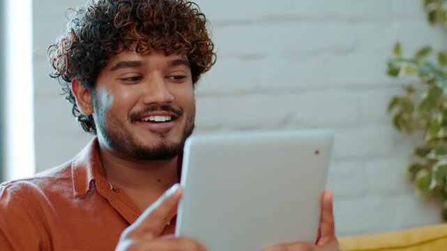 A young man with curly hair sits on a yellow couch, looking at a tablet. The room has a light-colored wall and plants in the background. He appears engaged and focused on what he is doing.