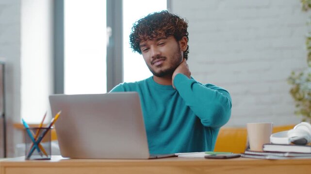 A young man with curly hair sits at a wooden desk in a bright room, looking at his laptop screen. He stretches his neck and appears to focus on his work. Various office supplies are nearby.