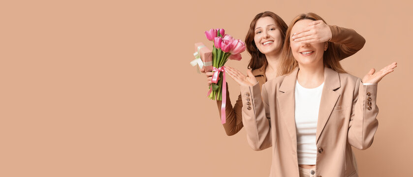 Beautiful mature woman and her daughter holding bouquet of tulips with gift box on brown background. Mother's Day celebration