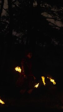Fire dancer performing dramatic nighttime routine with flaming batons and traditional costume in a dark forest setting, creating swirling trails of light and sparks