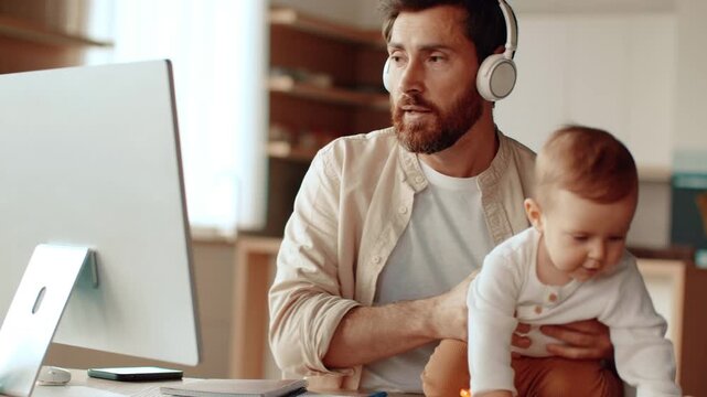 A man with a beard wearing headphones interacts with a baby during a work-from-home session. The bright indoor space features a desk and computer setup.