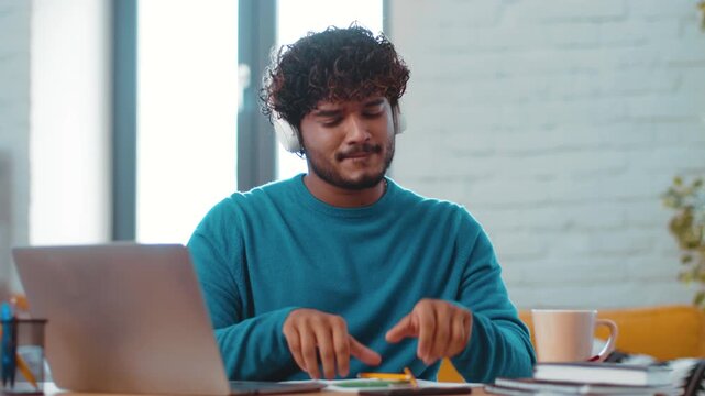 A young man with curly hair sits at a table in a bright room. He wears a blue sweater and enjoys music through headphones while working on a laptop. A cup and notebooks are on the table.